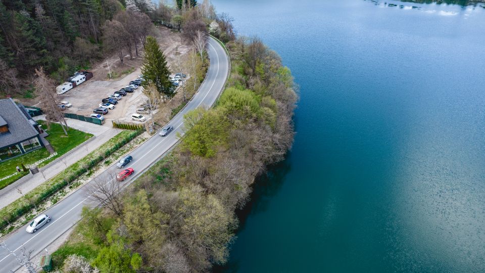 Въздушен изглед към извита двулентова пътна отсечка покрай брега на голямо синьо езеро. От едната страна има гориста ивица и малък паркинг до сграда, а от другата – спокойна водна повърхност по крайбрежието.