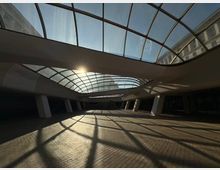 Покрита градска площ или вътрешен двор с извита стъклена конструкция на покрива и наклонени колони. Слънчевата светлина преминава през стъклата и хвърля решетъчни сенки върху павирания под.