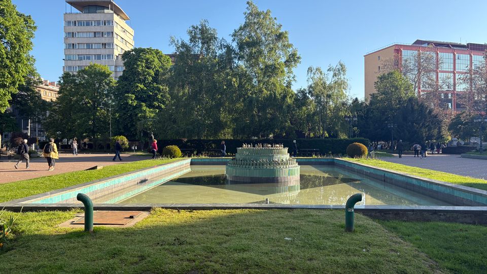 Градски парк с правоъгълен плитък басейн и декоративен фонтан в центъра, ограден от тревни площи и алеи. На заден план се виждат високи сгради, дървета и открито небе.