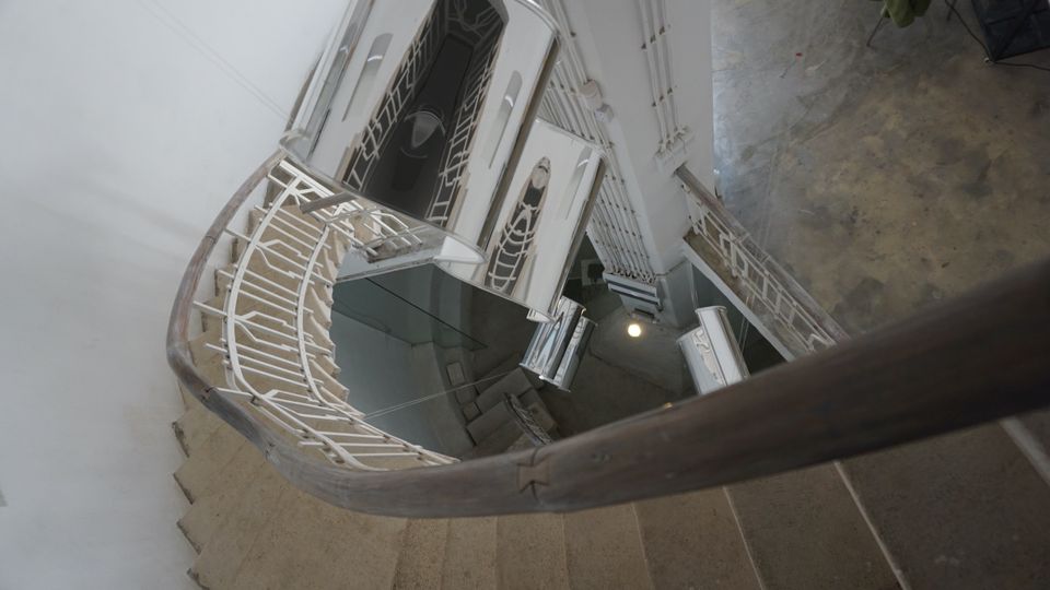 Поглед отгоре към вътрешно стълбище със завиващи стъпала и бели метални парапети с геометричен орнамент. В центъра се вижда шахта с асансьор и врати на различни етажи, а подът отгоре е бетонен.