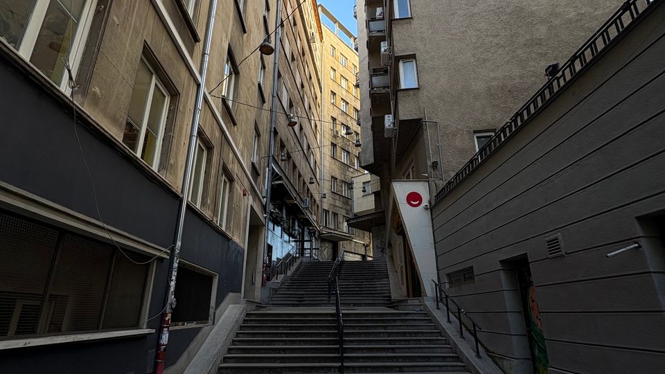 Тясна градска уличка с калдъръм, която води към широки стълби с парапети, разположени между високи жилищни сгради с прозорци и балкони. Над стълбите има опънати кабели и висящи улични лампи, а отгоре се вижда открито синьо небе.