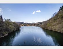 Спокойно езеро, разположено в долина между ниски гористи хълмове, с дървета по двата бряга и отражения във водата. На заден план се виждат далечни възвишения под ясно синьо небе с единични облаци.