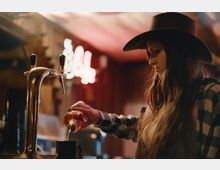 Интериор на бар с дървен плот и метална бирена колона с кранове на преден план. В дъното се вижда размазан неонов надпис и топло, приглушено осветление.