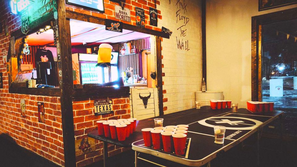 Интериор на бар с тухлени стени, неонови табели и винтидж плакати, с отвор към барплота и осветени хладилници. На преден план има маса за бирпонг с подредени червени пластмасови чаши, а вдясно се вижда входна врата с остъкление.