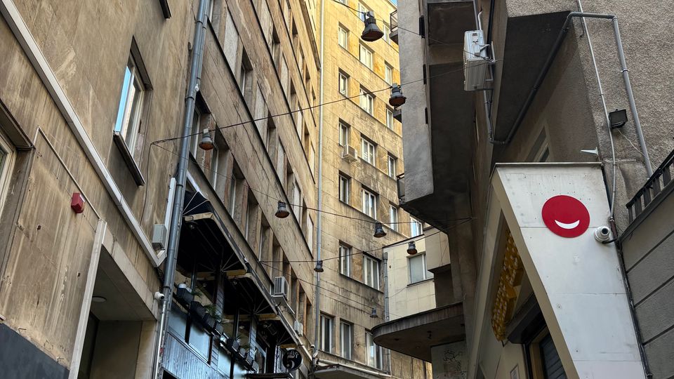 Тясна градска уличка между високи жилищни сгради, с широка стълба и метални парапети, водещи нагоре. Над стълбите има окачени улични лампи на кабели, а фасадите са с прозорци, балкони и външни климатични тела.