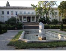 Бяла неокласическа обществена сграда с централен портик и колони, пред която има стълбище и три флагщока. На преден план се вижда правоъгълен каменен басейн с плитка вода и скулптура в средата, заобиколен от алеи и зеленина.