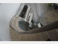 Поглед отгоре към вътрешно стълбище със завиващи стъпала и бели метални парапети с геометричен орнамент. В центъра се вижда шахта с асансьор и врати на различни етажи, а подът отгоре е бетонен.