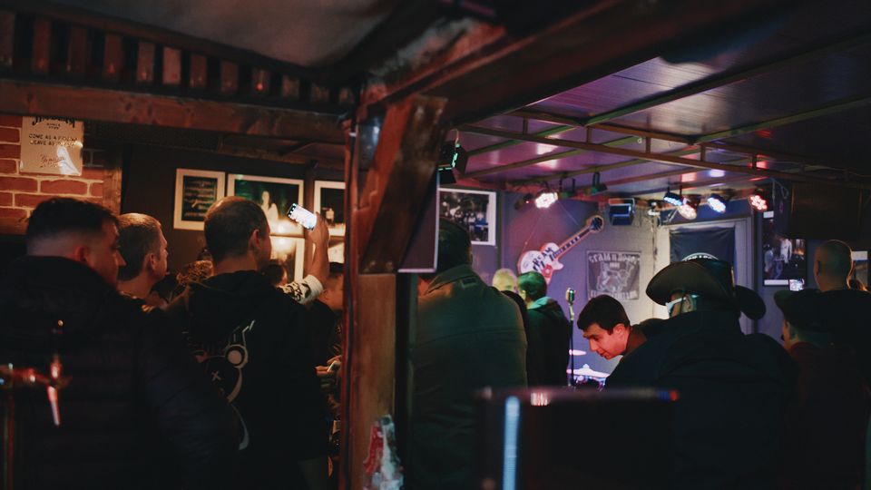 Интериор на малък музикален бар с тухлена стена, дървени греди и рамкирани снимки по стените. В дъното има ниска сцена с микрофон и сценично осветление, а над нея се виждат плакати и декоративна китара.