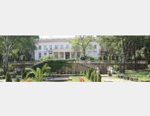 Бяла двуетажна неокласическа сграда с колонада и балкон, разположена зад терасирана градина. На преден план има каменни стълби и парапет, обрасли с бръшлян, алеи и цветни лехи сред високи дървета.