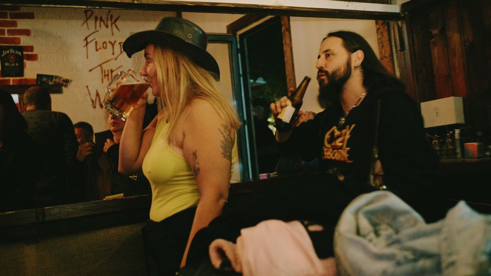 Интериор на малък бар с дървени греди и облицовка, отворена входна врата и барплот с бутилки и чаши. На стената има графит „Pink Floyd The Wall“ и частична тухлена декорация.